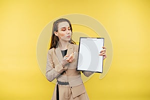 Business woman with a tablet in her hands stands on a yellow background looks at a blank white sheet of paper and shows a pen on