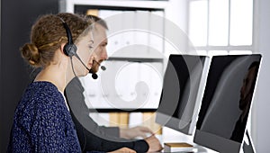 Business woman talking by headset while sitting with red-bearded colleague in modern office. Telemarketing and customer
