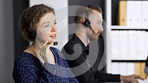 Business woman talking by headset while sitting with red-bearded colleague in modern office. Telemarketing and customer