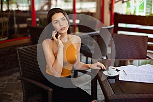 Business woman talking on the phone in a cafe at a table with scattered papers on the table and a cup of coffee