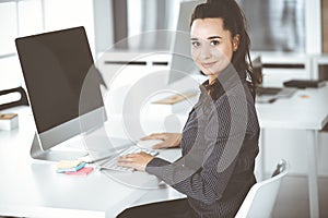 Business woman using computer at workplace in modern office. Brunette secretary or female lawyer smiling and looks happy