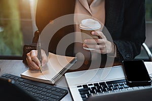 Business woman using laptop to work and writing on notebook with pen in office with mobile next to laptop