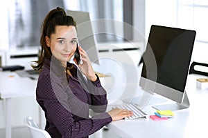 Business woman using phone and computer at workplace in modern office. Brunette secretary or female lawyer smiling and