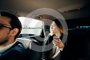 Business woman using smart phone and smiling while sitting on back seat in car