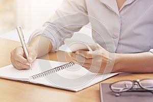 Business woman using smartphone and writing on notebook at the o