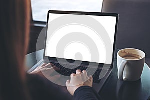 Business woman using and typing on laptop with blank white screen and coffee cup on table in cafe