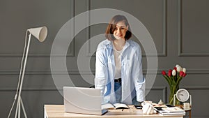 Business woman which looks straight ahead and which is wearing a white t-shirt, blue shirt and light jeans is standing near a desk