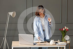 Business woman which is wearing a white t-shirt, blue shirt and light jeans is standing near a desk with a laptop and work