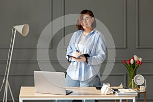 Business woman which is wearing a white t-shirt, blue shirt and light jeans is standing near a desk with a laptop and work