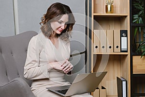 Business woman in white blouse sits on an armchair in her office with a laptop on her lap