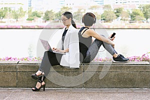 Business woman working on the laptop computer sitting near the relaxing sport woman in city park