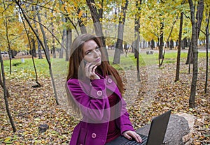 Business woman working with laptop and phone in autumn park. The woman has red hair and big green eyes.