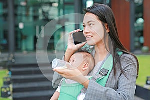 Business woman working by telephone with carrying her infant and feeding her child`s by milk bottle. Busy mother hurrying.