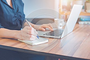 Business woman is writing on a notebook with a pen and using a laptop to work in the office