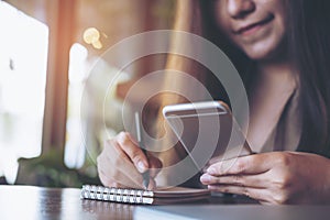 Business woman writing on notebook while using and holding mobile phone