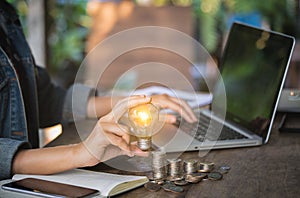 Business women hand holding light bulb on stack coins and working with computer on workplace.