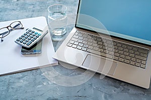 Business workspace with accounting  equipment like laptop, calculator, paper and water glass on a blue gray background, copy space