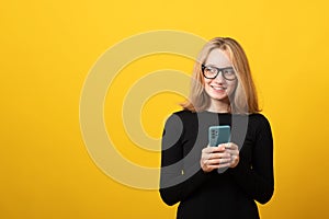 Business young blonde woman, in glasses, dressed in a strict black dress, holds a smartphone, is typing a text message. Copy space