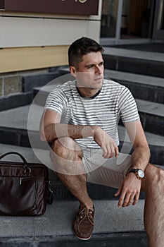 Business young man in a stylish summer striped T-shirt in fashionable shorts in suede shoes with a leather bag sits