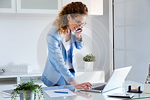 Business young woman talking on the mobile phone while using her laptop in the office.