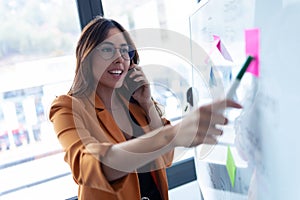 Business young woman talking on mobile phone while working on white board with post it stickers in the office