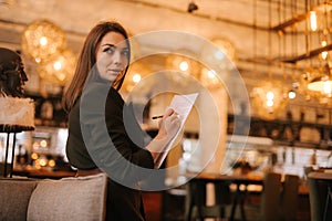 Business young woman wearing stylish clothes in luxury restaurant holding paper documents and pen.