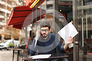 Businessman arguing by smartphone and reading papers at cafe tab