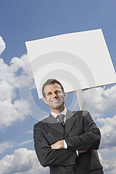 Businessman with arms crossed standing by blank sign against cloudy sky