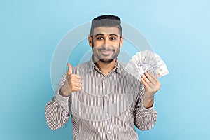 Businessman with beard holding dollar banknotes and showing thumbs up, enjoying rich life.