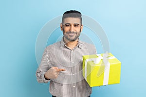 Businessman with beard standing, holding yellow gift box, pointing finger, looking at camera.