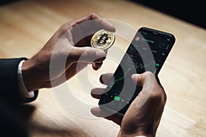 Businessman in black suit holding a golden bitcoin using Smartphone with Bitcoin trading chart on the screen to Trading