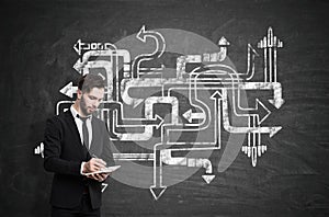 Businessman in a black suit with a notebook standing near a chalkboard with tangled arrows sketch on it.