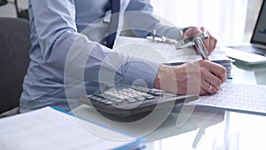 Businessman in blue t-short is using calculator and reviewing ring folder of financial documents with magnifying glass