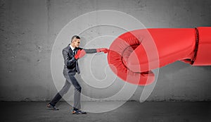 A businessman in boxing gloves on concrete background punches a giant red glove.