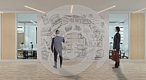 A businessman with a briefcase looks at the young man who draws a business sketch on the wall in a modern office