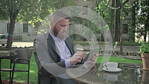 Businessman browsing tablet during coffe break. Outdoor. Steadicam shot. He is young and has beard. He is dressed on shirt and