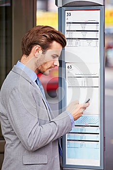 Businessman At Bus Stop With Mobile Phone Reading Timetable