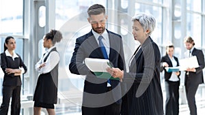 Businessman and businesswoman discussing work in office hall with colleagues on background