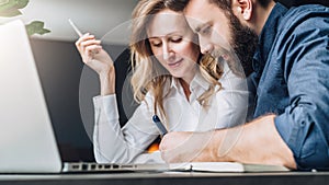 Businessman and businesswoman are sitting at table in front of laptop, discussing business concept. Man is writing pen