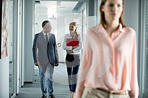 Businessman and businesswoman talking while walking in office corridor with female colleague in foreground