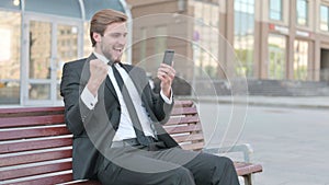 Businessman Celebrating Online Success on Smartphone, Sitting Outdoor on Bench