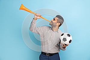 Businessman celebrating victory of favourite football team, blowing horn and holding ball in hands.