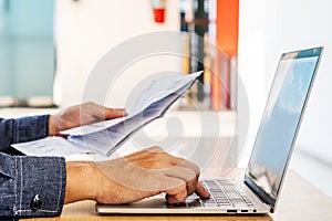 Businessman checking a typed report or document as he sits working on paperwork at a laptop computer, paying bills online on