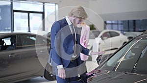 Businessman choosing the car in dealership close up. Tall man stands near automobile in auto show room, looking at car