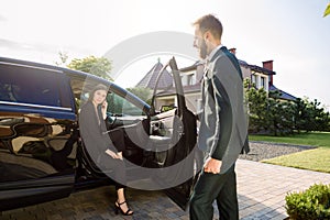 Businessman coming to car and opens door for young woman, talking phone. Businesswoman get out from the auto and is