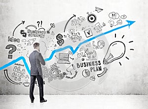 Businessman in a dark suit looking at a concrete wall with a growing blue graph and business plan icons depicted on it.