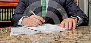 Businessman in dark suit sitting at office desk signing a contract with a library in the background.