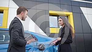 Businessman in a dealership shows a girl how to charge an electric car at a charging station