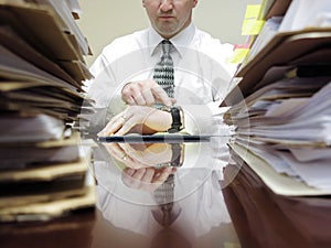 Businessman at Desk with Files Pointing at Watch