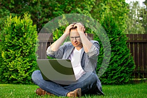 Businessman in desperation. Upset man sits on grass with laptop grabbing head